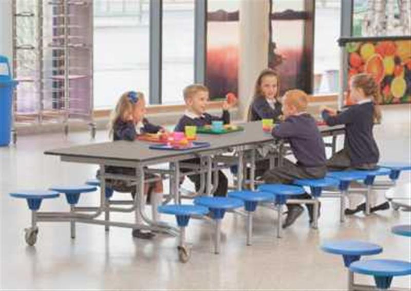 5 children are sat at a school dining table, which can seat a maximum of 16 pupils. The seats are blue and the table top is Grey Fleck, The table has been placed in a school dining hall.